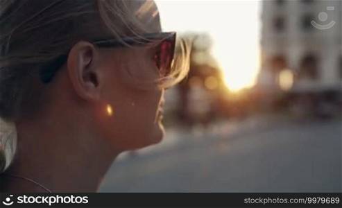 Close-up shot of a young woman in sunglasses looking into the distance during sunset, then she turns, smiles, and looks away again