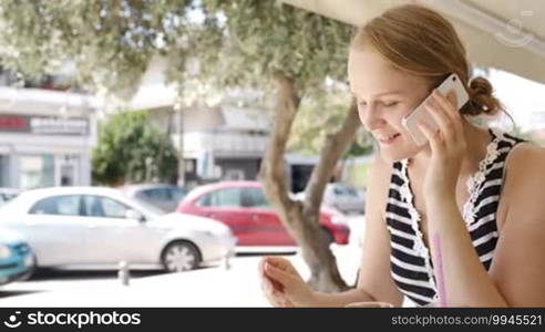 Close up shot of the attractive young woman sitting at an open-air cafeteria eating refreshments and using a mobile phone listening to the conversation as she eats