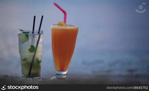 Close up shot of two fruity alcoholic cocktails - mojito and orange juice - standing side by side in their tall glasses in the sand on a tropical beach in the evening with copy space on the right