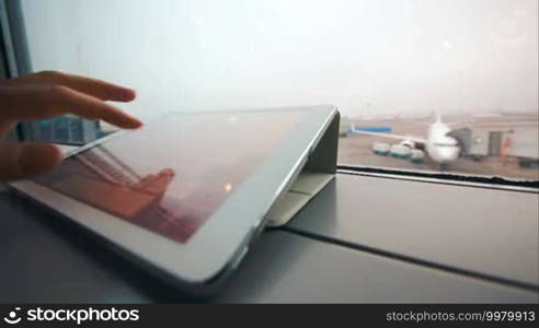 Close-up shot of woman hand typing on tablet PC by the window. Airport area and boarding plane in background