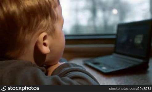 Close-up side view of a little boy watching film or cartoon on laptop while traveling by train. Focus on the boy