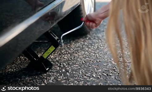 Close up view of woman's arm jacking up her car to change flat tire with spare. Young long-haired blonde is lifting up her car using a screw jack