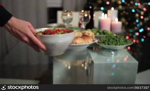 Close up woman's hand serving bowl of salad on festive table over blurred decorated Christmas table setting and Christmas tree background. Family celebrating Christmas Eve together at home.