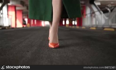 Closeup beautiful female wearing orange high heel shoes in emerald green coat walking in covered parking garage. Elegant woman's legs in high heel shoes walking alone in underground parking. Frontal view. Slow motion.