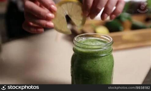 Closeup female hand decorating mason jar of fresh blended green smoothie with lemon slice and striped straw in the kitchen over blurry food ingredients on wooden tray background. Healthy vegetarian diet for weight loss and detox