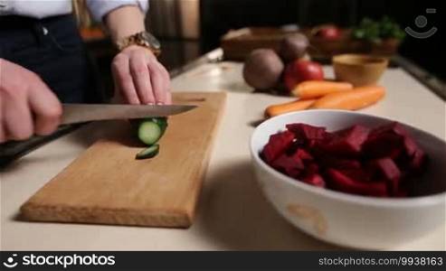 Closeup female hands with knife slicing cucumber on wooden chopping board in the kitchen while preparing healthy vegetable smoothie. Side view. Foreground bowl with sliced beet and vegetables on background.