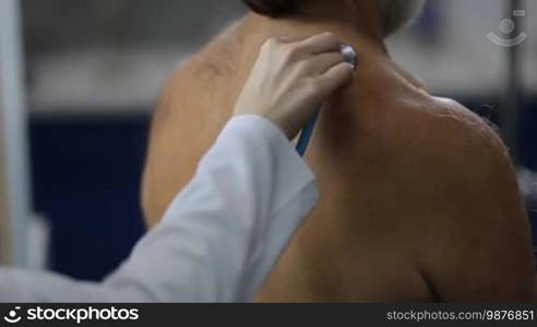 Closeup hand of female physician holding stethoscope and auscultating the lung sounds of senior ill male patient at hospital during medical checkup. Female doctor auscultating patient's back with stethoscope in clinic.