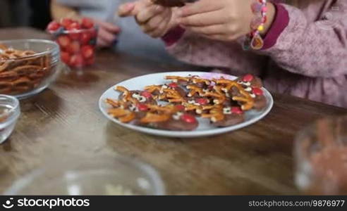 Closeup hands of multi-generation family taking Christmas decorated homemade cookies from a plate on a rustic wooden table background. Grandmother, daughter, and granddaughter eating tasty chocolate cookies in the kitchen. 180-degree dolly shot.