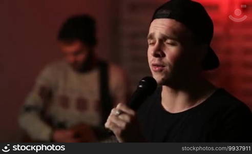 Closeup lead singer of music group holding microphone and singing. Blurred young musician playing acoustic guitar on background. Rock band performing on stage with spotlights.