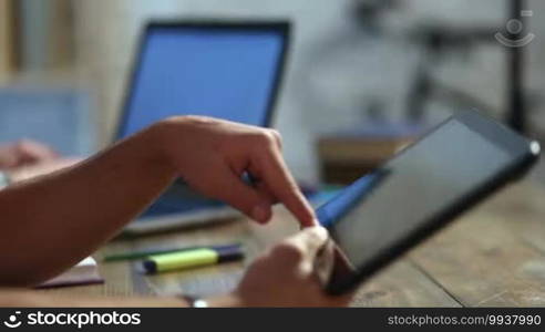 Closeup male student's hands using touchpad over blurred background with a friend working on laptop. Male freelancer working on digital tablet at home interior. Young teenage hipster scrolling and zooming on touchscreen device.