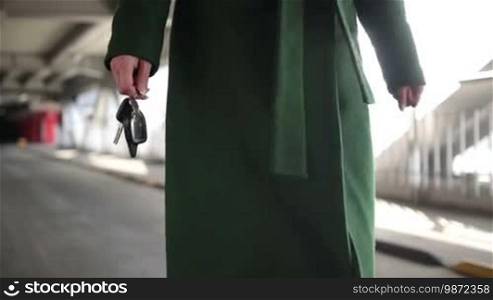 Closeup middle section of woman in emerald green coat with car key in hand walking to the car in covered parking garage. Female hand holding automobile key remote control. Focus on car key and the remote control car alarm systems. Slow motion