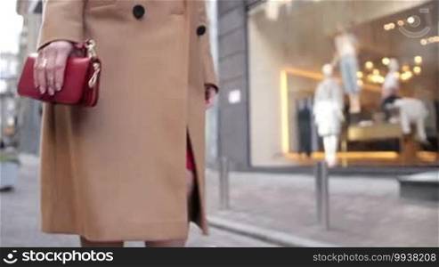 Closeup middle section of young female with red clutch wallet in her hand walking along city street for shopping. Young stylish woman wearing camel coat and red dress walking on sidewalk against showcase background. Slow motion.