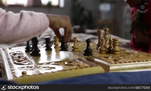 Closeup of a little girl playing chess, making a move by holding a black rook in her hand. Chess pieces are on the board. An African American child's hand moves the black rook two squares forward in the chess game while playing with her sister at home. Dolly shot. Slow motion.