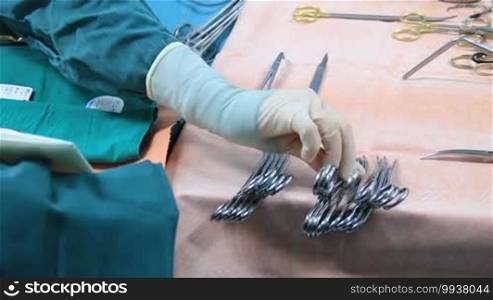 Closeup of gloved hand taking sterilized surgery instrument. Scrub nurse preparing surgical instruments for operation. Tools including scalpels, forceps, and tweezers arranged on a table for surgery.