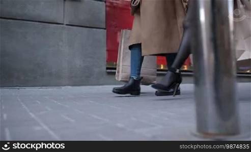 Closeup of slender female's legs in high heels and mini dress walking along the street against store showcase background. Slow motion. Side view. Beautiful fashionable woman carrying shopping bags going down cobblestone sidewalk after day shopping.