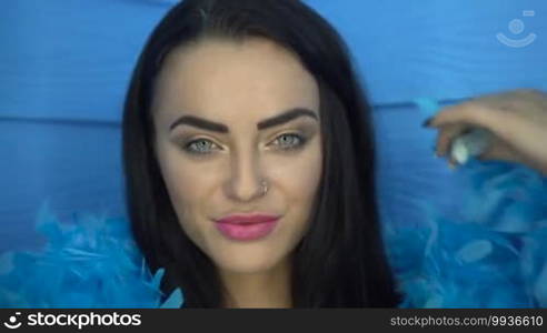 Closeup portrait of beautiful seductive brunette woman smiling and looking into the camera while playing with blue feather boa over blue wall background