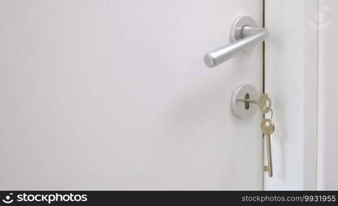 Closeup shot of door handle and keys in the door lock. Unseen woman in yellow and blue dress is opening the door and coming out of the room.