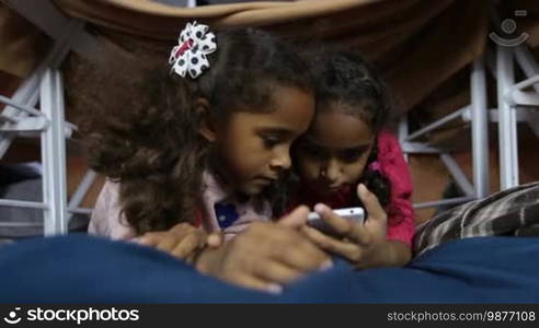 Closeup two adorable mixed race little sisters watching funny pictures on social network via smart phone as kids lie in cubby house made of chairs and blankets. Smiling African American girls playing online games on cellphone at home. Low view.