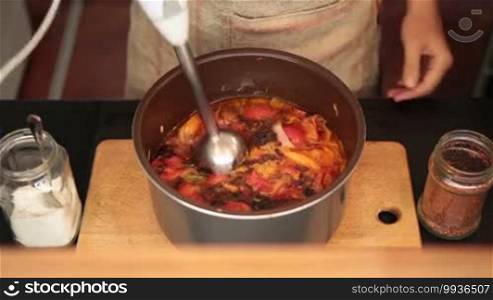 Closeup woman's hand mixing vegetable soup with blender in black pot on wooden board