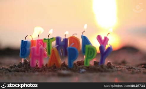 Colourful birthday candles spelling out the alphabet letters - Happy Birthday - standing upright in beach sand burning on a seashore at the edge of the sea