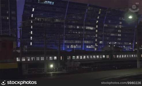 Commuter train passing by open platform in the city at night. Modern illuminated building in background