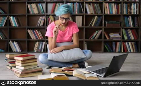 Concentrated college female student sitting on the floor, surrounded by a heap of books, researching with a laptop in the university library. Serious hipster girl with blue hair and trendy eyewear studying in the library with a laptop on the internet.