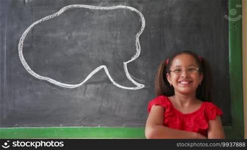 Concept on blackboard at school. Young people, student and pupil in classroom. Happy and funny latina girl in class. Portrait of female child smiling, looking at camera, saying words