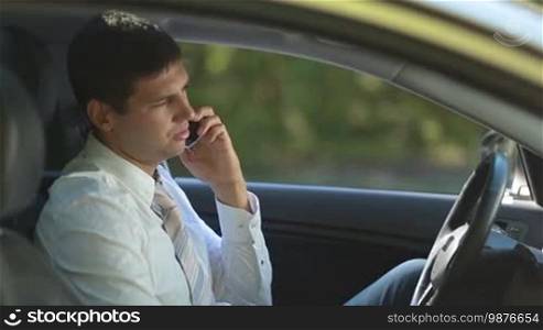 Confident businessman in necktie communicating on smartphone while sitting in car. View from car window. Side view. Handsome entrepreneur discussing business matter with his colleague on mobile phone while sitting in driver's seat.