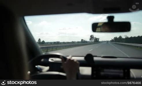 Confident woman driving car on motorway during summer road trip with beautiful landscape in the background. View from inside out of vehicle's interior.