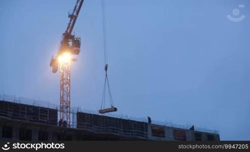 Construction works in the evening: crane lifting cargo. Winter snowing