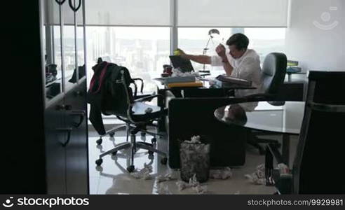 Corporate manager in a modern office tries to write a job letter. The man is frustrated and keeps on crumpling up paper. He plays with an anti-stress ball and touches his head with a desperate expression. Wide-angle shot