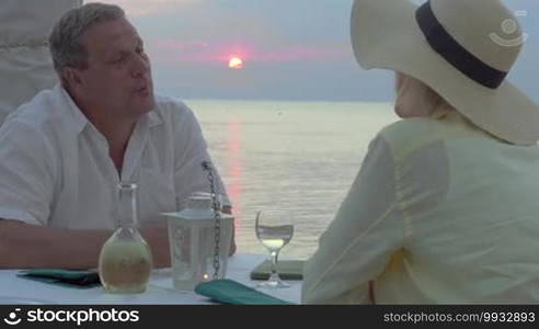 Couple is having a date in an outdoor restaurant at sunset, man is proposing a toast, they clink wine glasses.