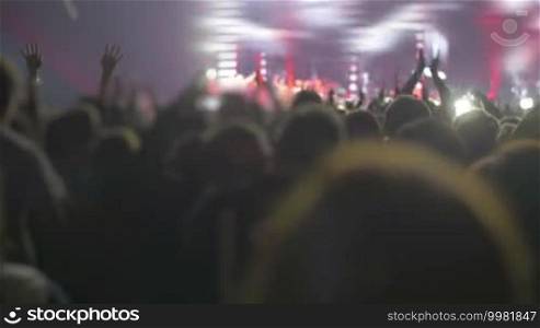 Crowd applauding during rock or pop concert in front of illuminated colored stage with huge screen showing singer