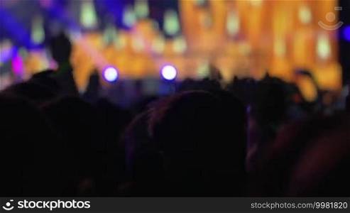 Crowd of fans at the concert. Favorite music rhythms making them feel the energy and dance. Illuminated colorful stage in the background