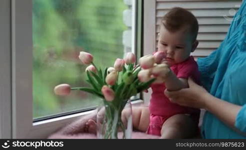 Cute infant child sitting on window sill and playing with tulip flowers. Adorable baby girl trying to bite tulip. Young mother holding little kid and showing flowers to the child while relaxing together at home.