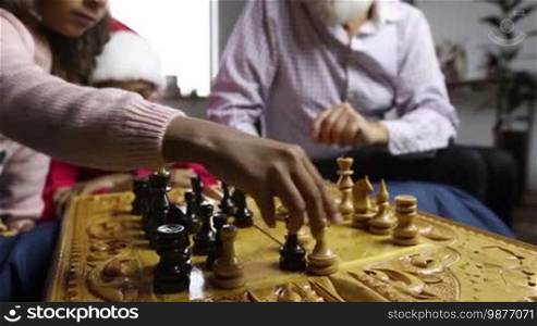 Cute little child capturing white pawn while playing chess game with her grandfather on Christmas. Happy mixed-race girl in Santa hat watching chess match between her little sister and grandpa at home. Focus on chessboard and players' hands. Dolly shot.