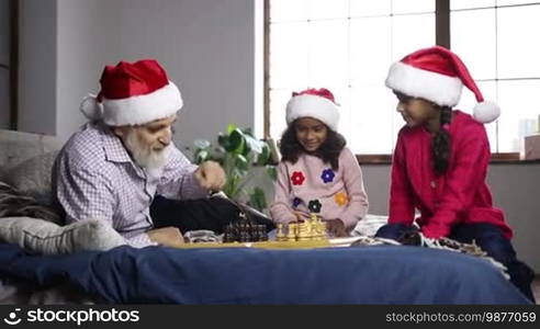 Cute mixed race girl in Santa hat playing chess with grandfather while sitting on bed at home. Smiling little girl learning chess game with granddad and making moves as excited younger sister clapping hands while family spending leisure on Xmas eve.