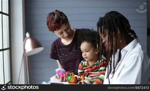 Cute mixed race toddler boy having fun with parents in bedroom at home. Interracial family lounging together. Handsome African father with dreadlocks and cheerful Caucasian mother playing together with son with toys while child sitting on dad's lap.
