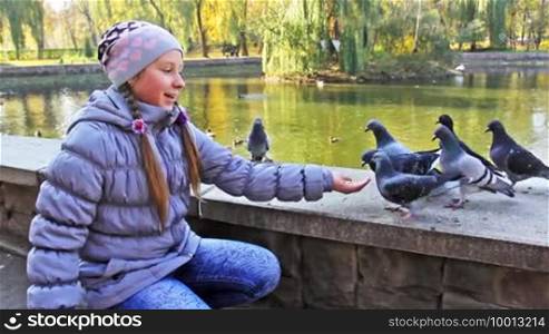 Cute teen girl sitting feeds popcorn to pigeons from hand in autumn city park near lake, closeup