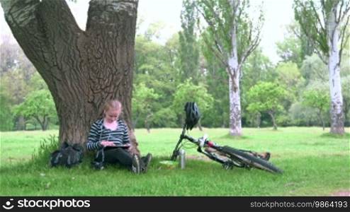 Cyclist girl sitting under tree in the park, listening to music