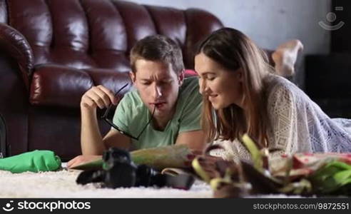 Delighted couple lying barefoot on the floor at home and planning a travel with paper map. Smiling charming brunette woman and positive man reading a paper map and discussing future journey while preparing for holiday vacation trip in living room.