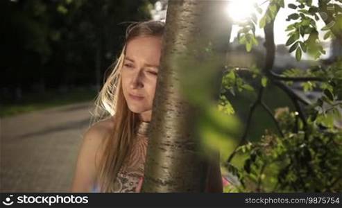 Disappointed woman leaning her head on a tree trunk in the park. Depressed girl with eyes full of sorrow, almost crying, standing alone near a tree outdoors. Beautiful woman experiencing emotional stress and depression. Slow motion. Steadicam stabilized shot