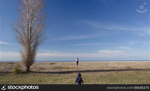 Distant view of a little boy running to his mother. Scenic landscape around with a lonely tree, blue sky, and sea