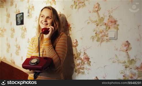 Dolly shot of a smiling young woman having a phone talk at home. She is standing leaning on the wall
