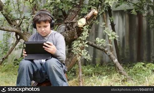 Dolly shot of a teenager boy sitting outdoors in headphones and using a tablet computer