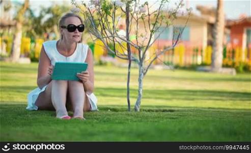 Dolly shot of beautiful woman wearing sunglasses sitting barefoot on a lawn in the shade of a tree with a laptop computer on her lap