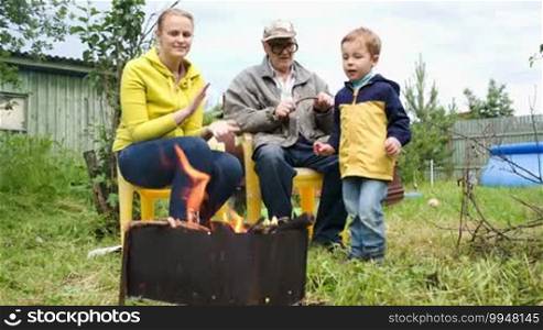 Dolly shot of mother, son, and grandfather by the fire in the yard. Boy throwing firewood into the fire, mother applauding