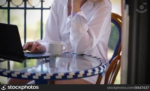 Dolly shot of pretty cheerful woman using her computer, having tea and focusing on her phone conversation