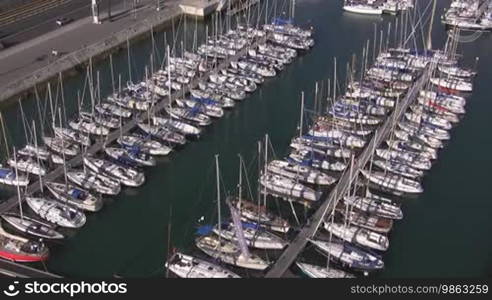 Ein Yachthafen; an Docks angelegte weiße Segelboote liegen in einem Hafen nebeneinander. Direkt am Hafen eine befahrene Straße.
Formatted (Translated):
A marina; white sailboats moored at docks are lined up next to each other in a harbor. A busy road runs directly by the harbor.