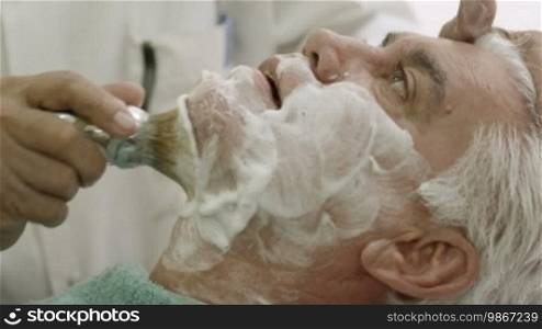 Elderly barber working in old style shop or men's beauty parlor and shaving client with razor